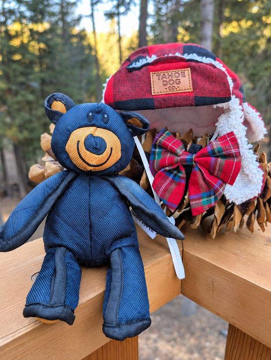 Red plaid dog trapper hat, red tartan bow tie, and Tahoe bear toy displayed on a wooden railing with pinecones.