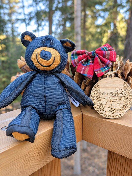 Tahoe bear dog toy, red tartan bow tie, and wooden Tahoe Dog ornament displayed on a deck railing.