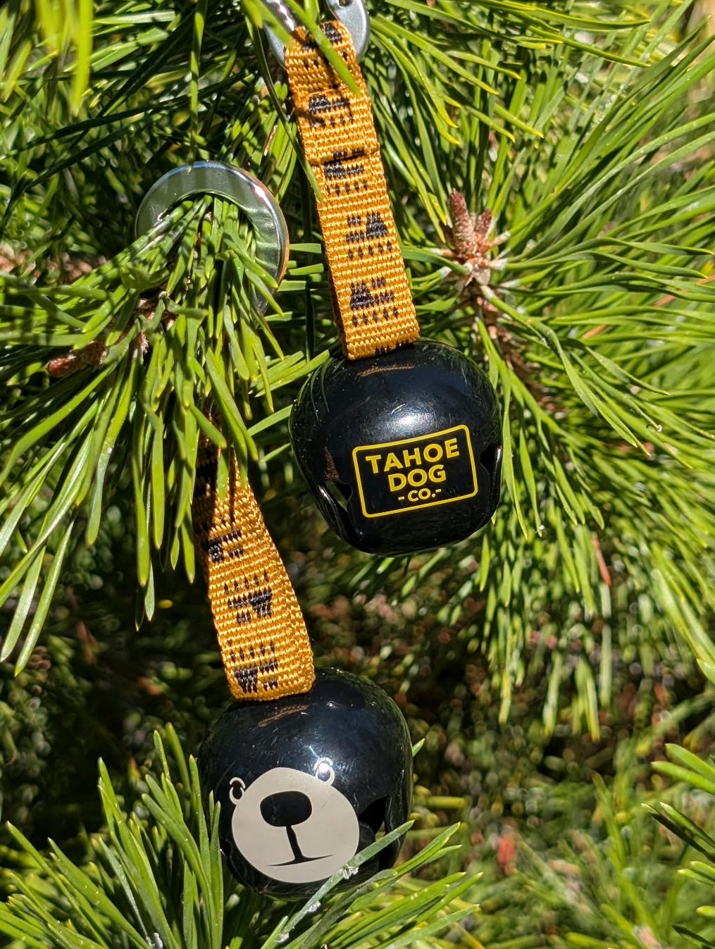 Two black trail bells with yellow straps, showing Tahoe Dog Co. logo and bear face designs among pine branches.