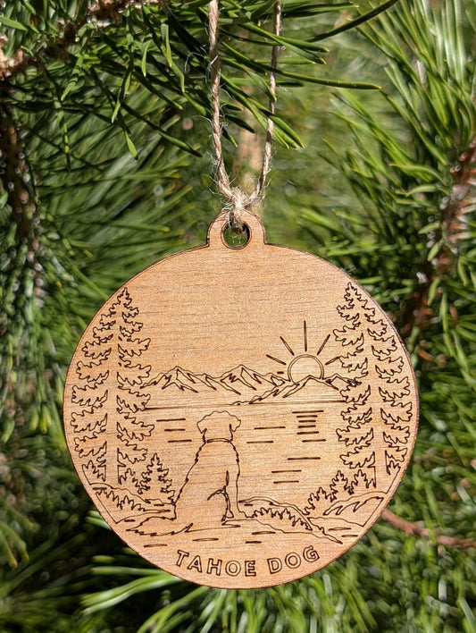 Round wooden Tahoe Dog ornament engraved with a dog overlooking a lake and mountains, hanging from a pine branch.